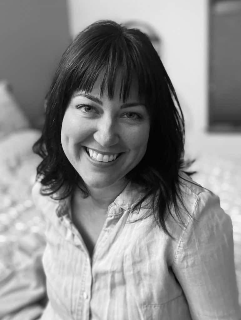 photo of woman smiling at camera with head slightly tilted to her left. She is wearing a denim button down shirt and the photo is in grayscale